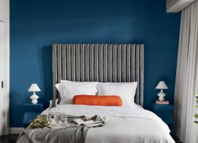 A bedroom, containing a bed with a large headboard against a blue wall, flanked on both sides by blue nightstands